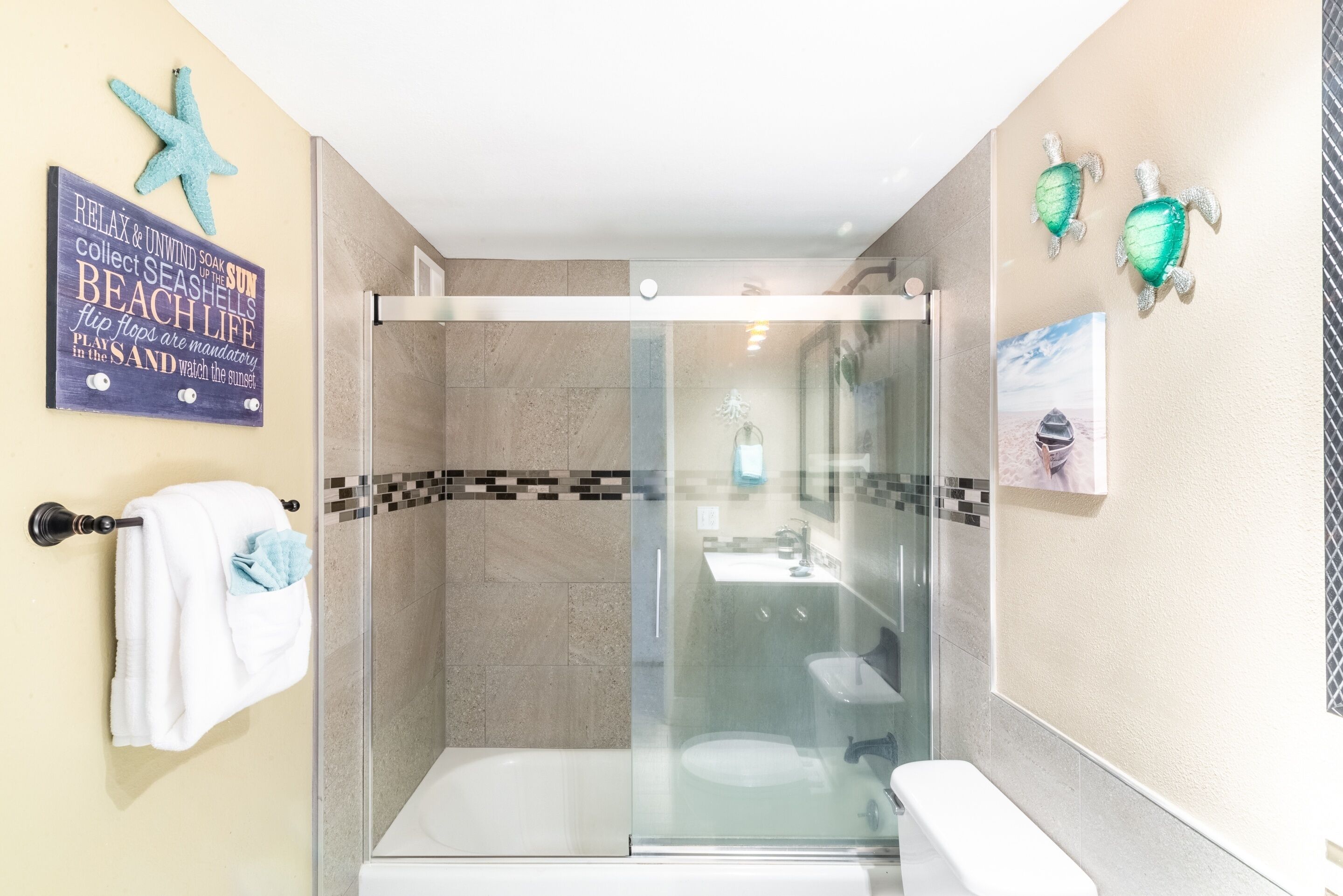 Ein Badezimmer mit einer gläsernen Duschkabine, einer weißen Toilette und einem Waschbecken. Ein Handtuchhalter mit Handtuch und eine Meerstern-Dekoration sind an der Wand angebracht.