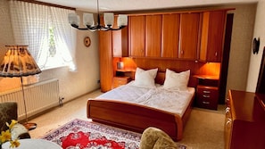 A bedroom with a wooden bed, bedside lamps, a radiator, a window with curtains, and a patterned rug.