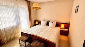 A hotel room with a large bed, a bedside table with a lamp, a bench at the foot of the bed, and a window with curtains. - "Auf dem Heidchen" Vacation Home, on the edge of the forest, with a sauna and a large garden (Knüllwald)
