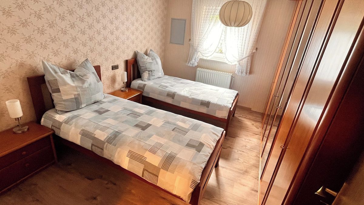 Ein Schlafzimmer mit zwei Betten, Holzboden, einem Fenster mit Vorhängen und einem Heizkörper.