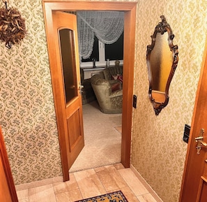 A hallway with patterned wallpaper, a wooden door, a mirror, and a glimpse of a room with a couch and a window with curtains. - "Auf dem Heidchen" Vacation Home, on the edge of the forest, with a sauna and a large garden (Knüllwald)