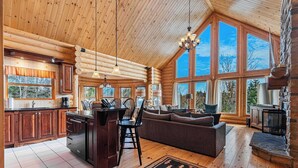 A log cabin interior with a kitchen, dining area, and living room, featuring large windows and wooden ceilings. - Spa, Pool & Sauna | Sérénité des Laurentides (Mille-Isles)