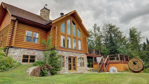 A log cabin with a stone foundation, large windows, and a wooden deck. - Spa, Pool & Sauna | Sérénité des Laurentides (Mille-Isles)