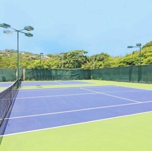 Ein Tennisplatz mit Netz, umgeben von grünen Zäunen und Bäumen.