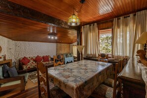 Um quarto aconchegante com teto de madeira, uma mesa de jantar com toalha de mesa estampada, um sofá com almofadas vermelhas e uma janela com cortinas.