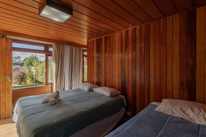 Quarto com painéis de madeira, duas camas, janela com cortinas e vista para o verde lá fora.