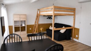 Un lit superposé avec une échelle, un canapé noir, un meuble en bois et une table à manger avec des chaises.