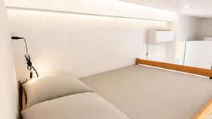 Un lit simple avec un cadre en bois et une tête de lit, une table de chevet avec une lampe, et un mur blanc agrémenté d’un luminaire au plafond.