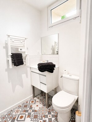 Un baño con inodoro blanco, lavamanos con espejo, toallero y baldosas de suelo estampadas.