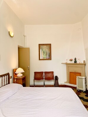 A bedroom with a bed, a nightstand with a lamp, a chair, a fireplace, and a framed picture on the wall. - Natura tra bosco e lago (Castelveccana)