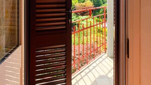 Un balcó amb barana vermella, una porta amb llistons horitzontals i vista a vegetació i muntanyes.