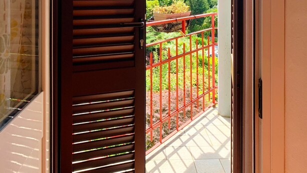 Un balcon avec une balustrade rouge, une porte à lattes horizontales et une vue sur la verdure et les montagnes.