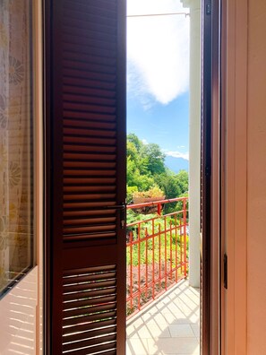Ein Balkon mit roter Brüstung, eine Tür mit Rollläden und Blick auf Grünflächen und Berge.