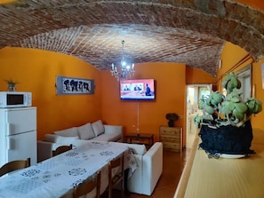 A room with a brick ceiling, orange walls, a television, a dining table with a patterned tablecloth, a sofa, and a potted plant.