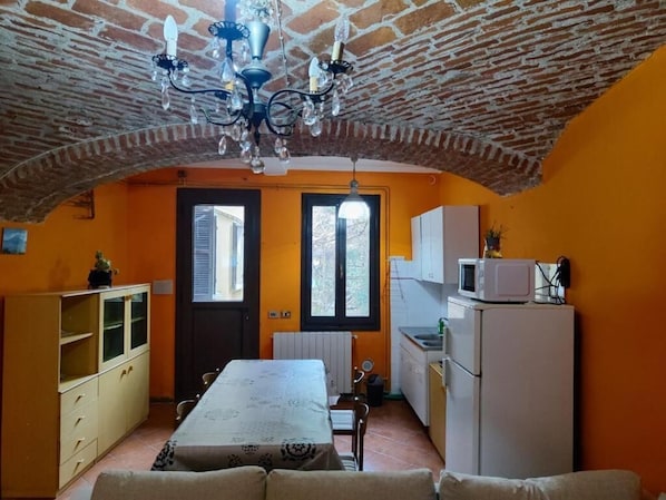 A room with a brick ceiling, a dining table, a kitchen area with a refrigerator, and a window with shutters.