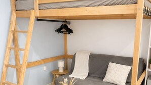 Een stapelbed met een ladder, een grijze bank, een klein rond tafeltje met vazen, en een witte stoel.