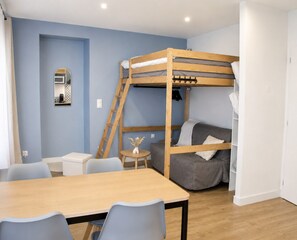 Una cama litera con escalera, una mesita redonda con un florero, un sofá gris, una mesa de madera con sillas y una puerta blanca.