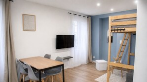 Een kamer met een houten tafel, stoelen, een televisie, een stapelbed en een klein wit kastje.