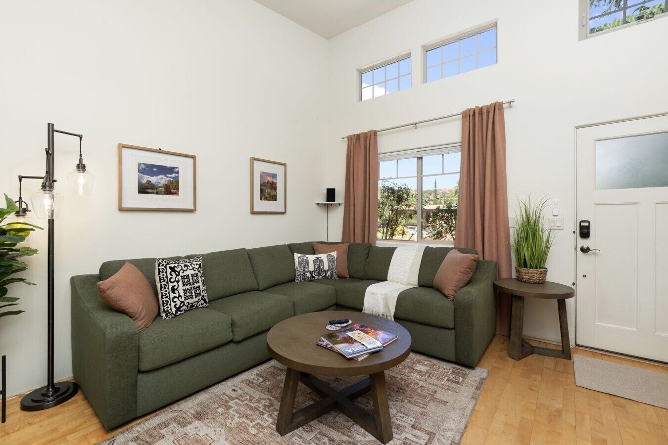 A living room with a green sofa, a coffee table, a floor lamp, framed pictures on the wall, a window with curtains, and a door with a plant beside it.