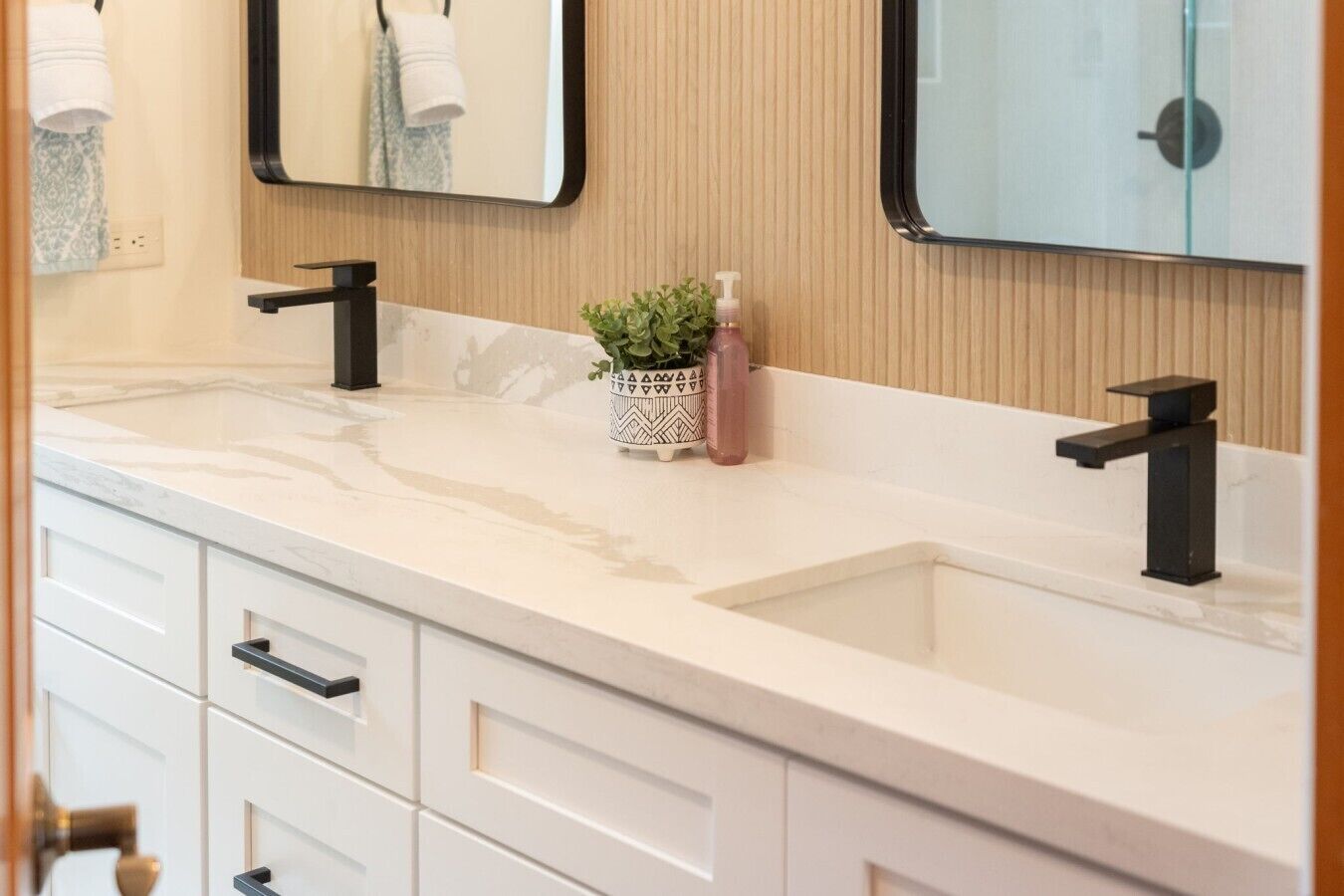 Une salle de bain avec un comptoir en marbre blanc, deux robinets noirs, une petite plante en pot et un miroir sur lequel est accrochée une serviette.