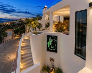 Ein modernes Gebäude mit einer Treppe, die zu einem Balkon führt, einem Schild mit einem Picasso Gemälde und Blick auf eine Küstenstadt.
