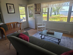 Ein Zimmer mit einem grünen Sofa, einer Holzbank, einem weißen Stuhl, einem kleinen Tisch mit einer Schachtel und Blick auf einen See durch große Fenster.