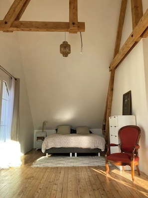 Ein Schlafzimmer mit schrägen Wänden, Holzbalken, einem Bett mit gemustertem Überwurf, einem roten Sessel und einem Nachttisch.