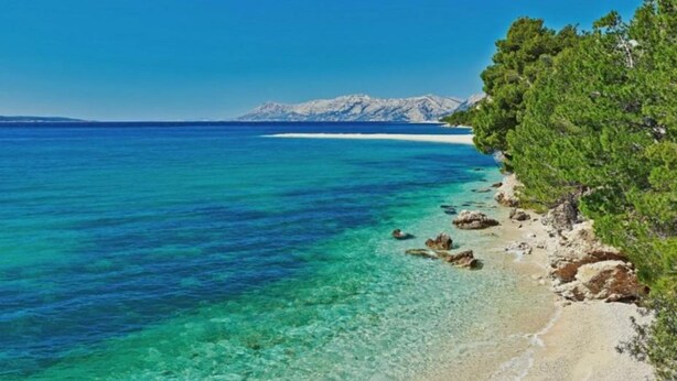 Una playa con agua turquesa cristalina, una orilla arenosa y una cadena montañosa al fondo.