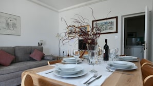 Comedor con mesa de madera dispuesta para seis personas, rodeado de sillas, un jarrón con ramas y una botella de vino sobre la mesa.