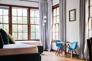 Ein Zimmer mit großen Fenstern, einem Bett, einem Tisch mit Stühlen und einer Stehlampe.