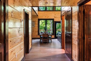 A wooden interior hallway leading to a dining area with a table and chairs, and a view of the outdoors through large windows. - Mountain Laurel Cabin!! Heated Pool, Theatre, Hot-tub, Games, near Pigeon Forge! (Sevierville)