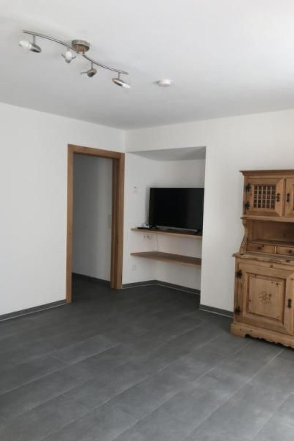 Una stanza con pavimento piastrellato grigio scuro, un armadietto in legno, un televisore su uno scaffale e un lampadario.