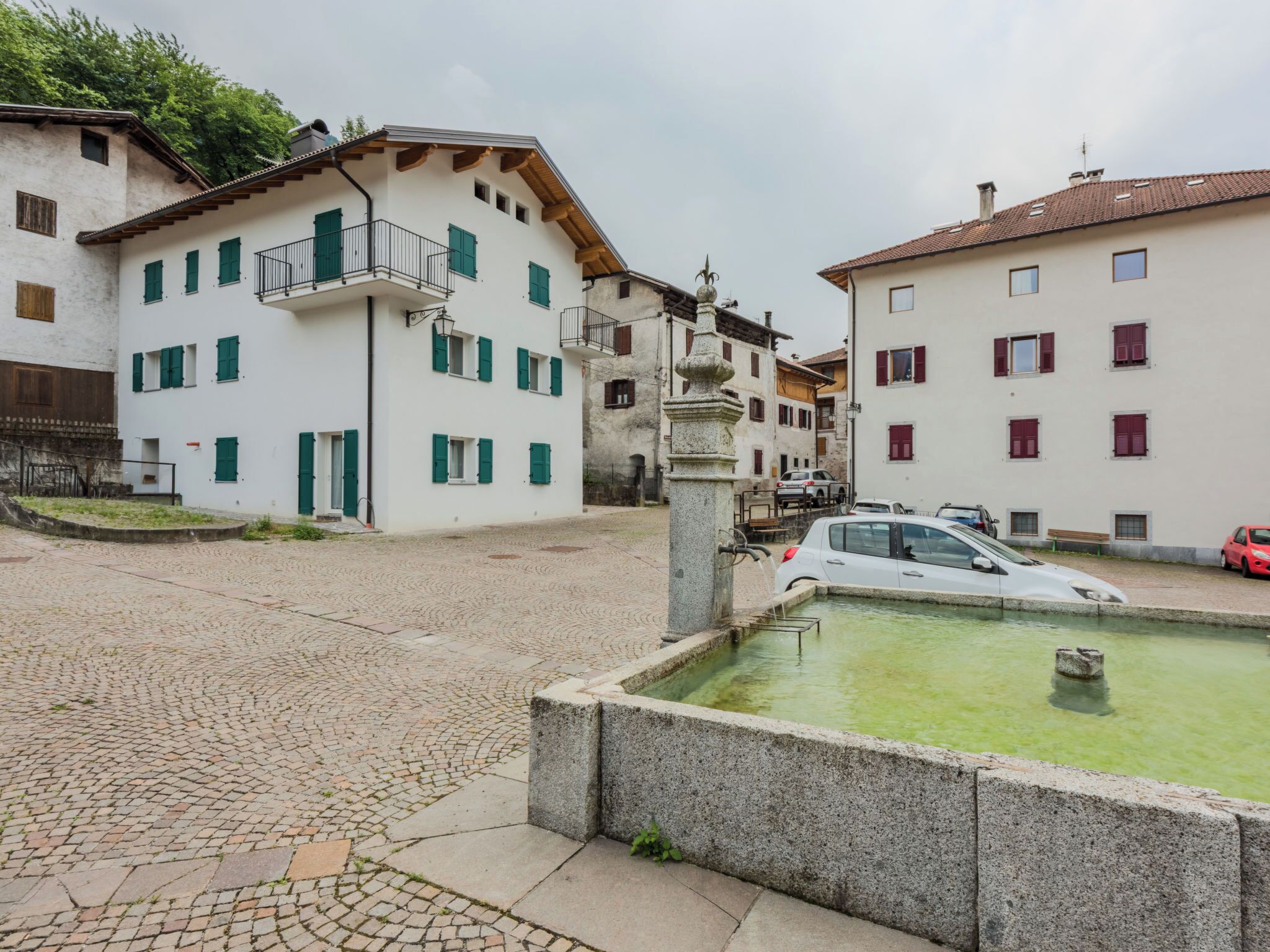 A cobblestone street with buildings, a fountain, and parked cars.
