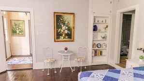 A bedroom with a bed, two chairs, a small table, a bookshelf, and a painting on the wall.