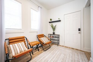 Ein Zimmer mit zwei Sessel, einem kleinen Tisch mit Topfpflanze, einer weißen Tür und einem fenster mit Vorhängen.