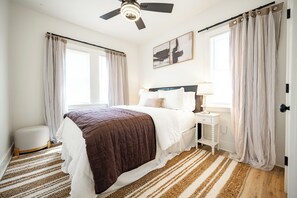 Ein Schlafzimmer mit Bett, Nachttisch, Lampe, Vorhängen und Deckenventilator.