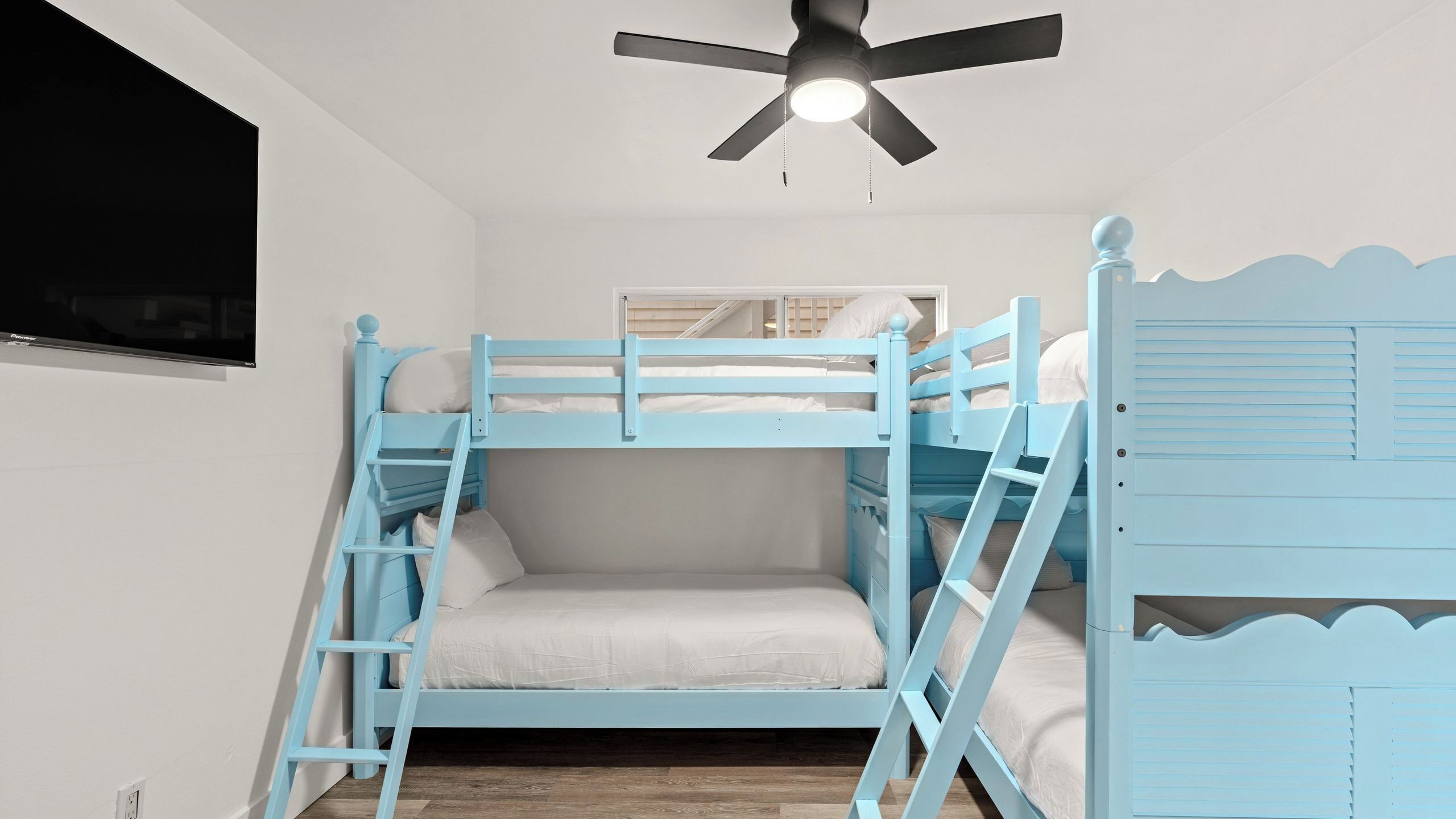A bunk bed room with a ceiling fan, a TV mounted on the wall, and a wooden floor.