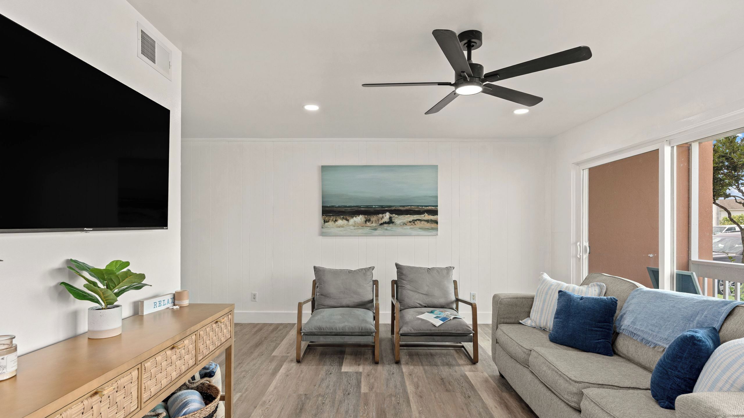 Un salon équipé d’un téléviseur à écran plat, d’un ventilateur de plafond, de deux fauteuils gris, d’un canapé gris et d’un tableau représentant une scène de plage.