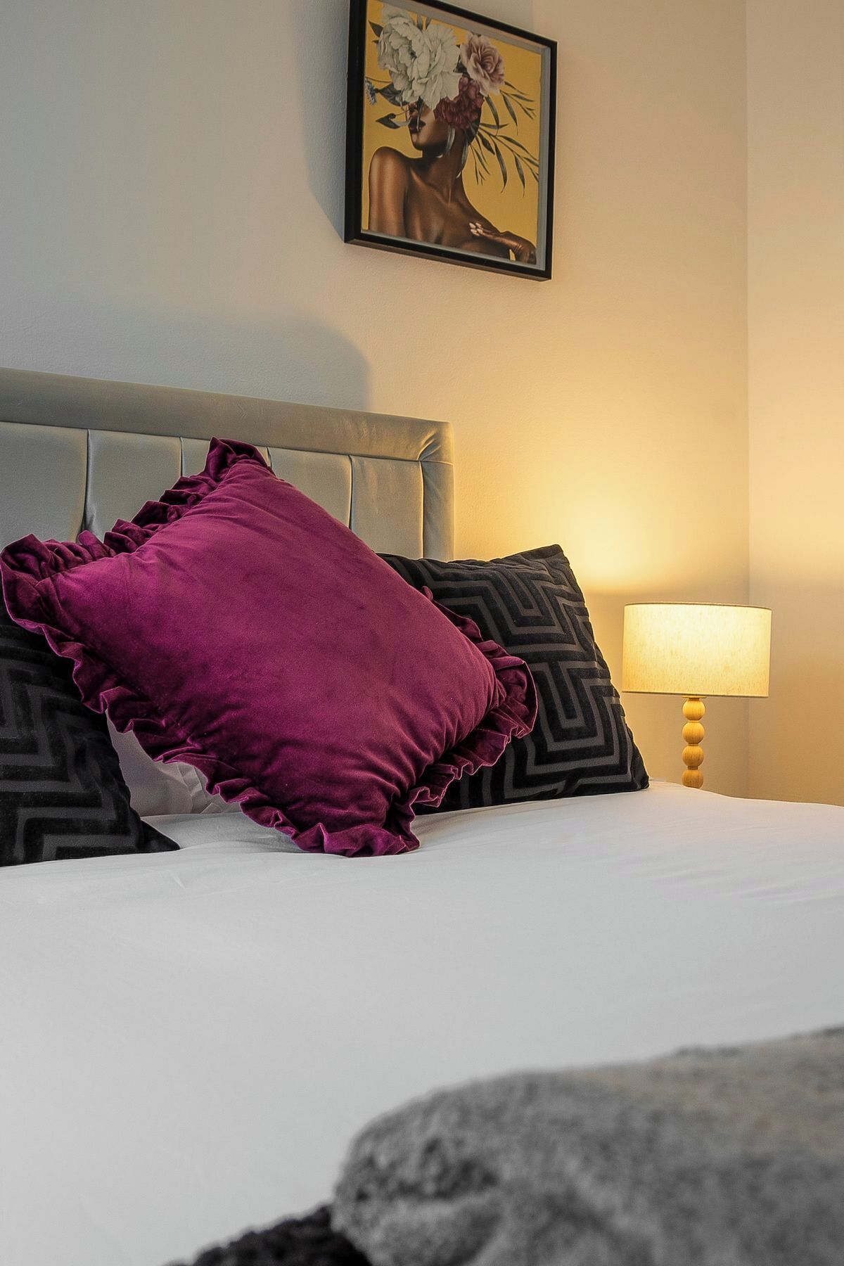 A bed with a ruffled purple pillow, a patterned pillow, and a wooden bedside table with a lamp.