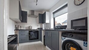 A compact kitchen with grey cabinets, a built-in oven, a microwave, and a washing machine. - The Rushden Retreat • 1-Bed Flat + Parking (Rushden)