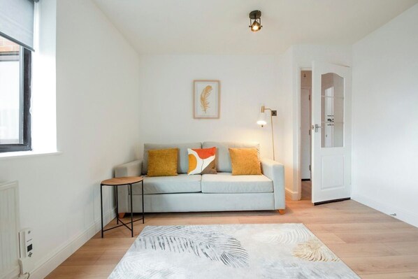 A living room with a light grey sofa, a small side table, a patterned rug, a framed artwork on the wall, and a door leading to another room.