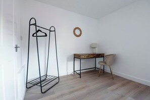 A room with a metal clothes rack, a wooden desk, a chair, and a wall-mounted lamp.