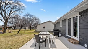 Un patio con mesa y sillas, una parrilla y una casa con ventanas amplias.
