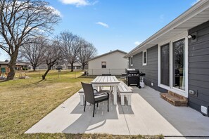 A patio with a table and chairs, a grill, and a house with large windows.