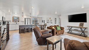Una cocina moderna con armarios blancos, un comedor con mesa y sillas, y una sala de estar con sofás de cuero y televisión.