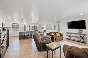 A modern kitchen with white cabinets, a dining area with a table and chairs, a living room with leather sofas and a TV.