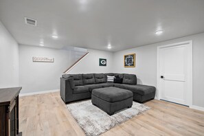 Un salon moderne avec un canapé d’angle, un pouf gris, un plancher en bois et une porte blanche.