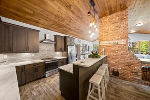 A modern kitchen with dark wooden cabinets, a large island with bar stools, and a brick accent wall. - Summit View Ski and Golf Retreat w/hot tub & Sauna (Winhall)