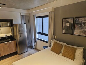Hotellihuone, jossa on sänky, pieni keittiönurkkaus, ikkuna verhoilla ja taideteoksia seinällä.