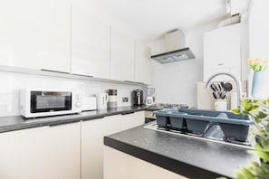 A modern kitchen with white cabinets, a black countertop, a microwave, a coffee maker, a kettle, a toaster, a sink, and a vase with flowers.